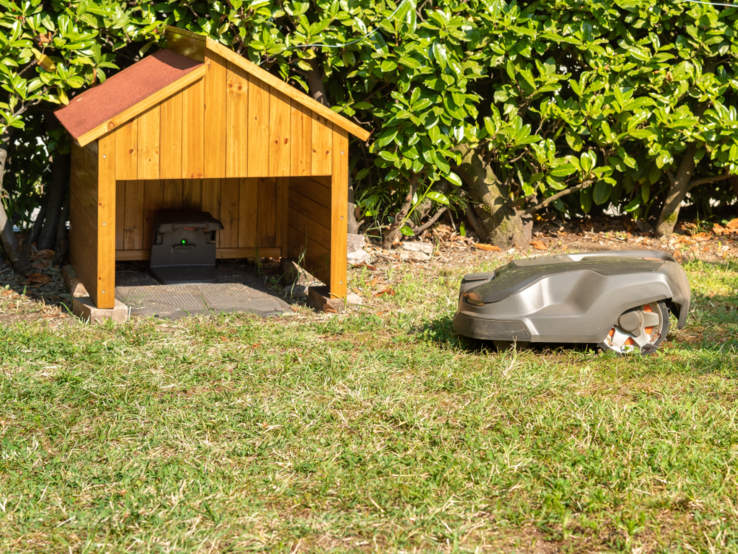 Mähroboter Garage Ein Rasenmähroboter fährt bei Sonnenschein über eine Wiese in Richtung einer kleinen DIY-Garage mit Ladestation.
