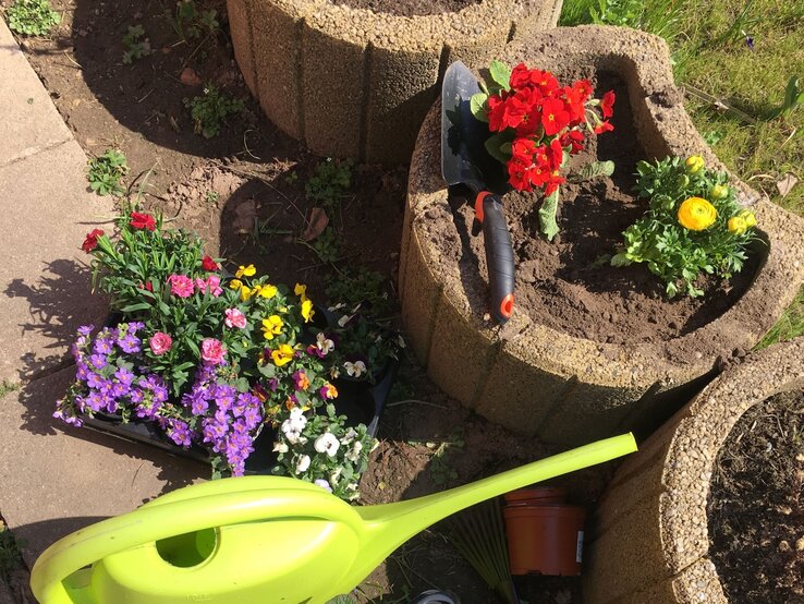 Zwei bepflanzte Pflanzsteine mit bunten Blumen, daneben liegen eine grüne Gießkanne, Blumenkelle und eine Pflanzenpalette auf sonnigem Boden.