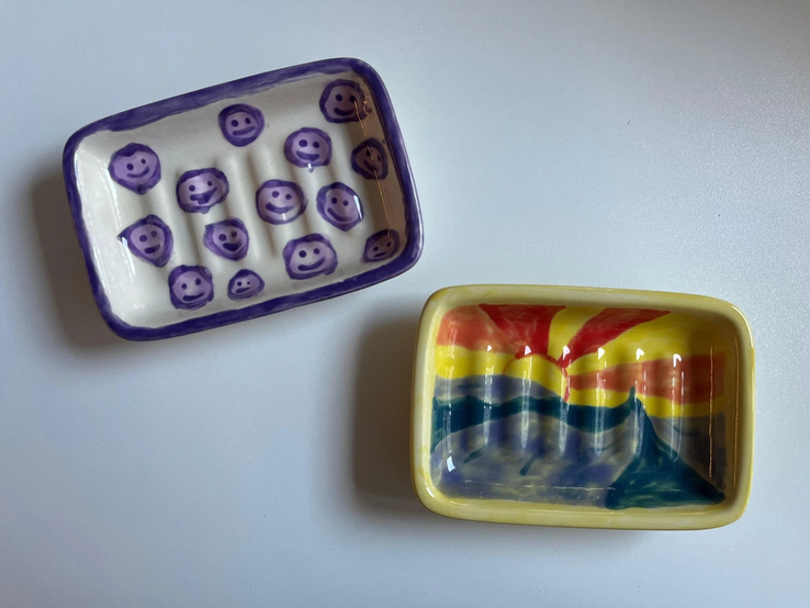 Zwei bemalte Keramikschalen, eine mit lila Smileys und die andere mit einem farbenfrohen Sonnenaufgang-Motiv, liegen auf weißer Oberfläche.