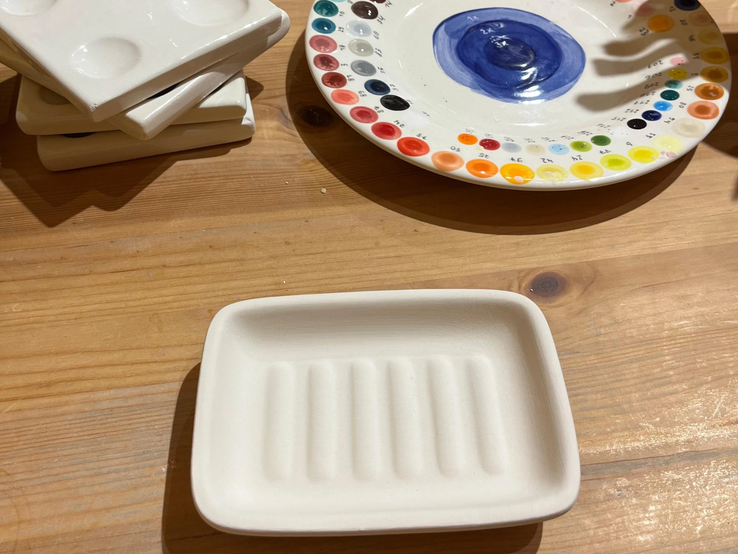 Eine unbemalte Keramikschale mit Rillen liegt neben Farbtellern und stapelbaren Paletten auf einem Holztisch.