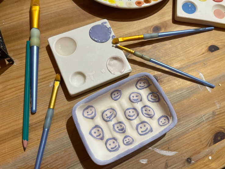 Bunte Farbpinsel und Paletten liegen neben einer Keramikschale mit lila Smileys, arrangiert auf einem Holztisch.