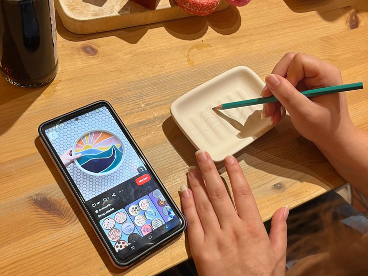 Seifenschale bemalen Vorbild Skizze | © Kaja Wundersitz/ Linda Pötzsch/ BIld der Frau Eine Hand zeichnet mit einem grünen Stift ein Wellenmotiv in eine Keramikschale, daneben ein Smartphone mit ähnlicher Vorlage. | © Kaja Wundersitz/ Linda Pötzsch/ BIld der Frau