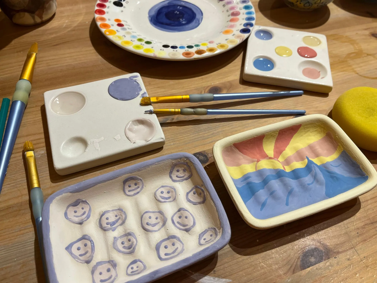 Seifenschale bemalen DIY | © Kaja Wundersitz/ Linda Pötzsch/ BIld der Frau Zwei bemalte Keramikschalen mit Smileys und Sonnenmotiv liegen neben Farbtellern, Pinseln und einem gelben Schwamm auf Holztisch. | © Kaja Wundersitz/ Linda Pötzsch/ BIld der Frau