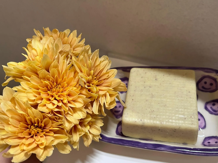 Ein Stück beigefarbene Seife mit feiner Struktur liegt in einer lila-weiß gemusterten Schale, daneben gelbe Chrysanthemen.