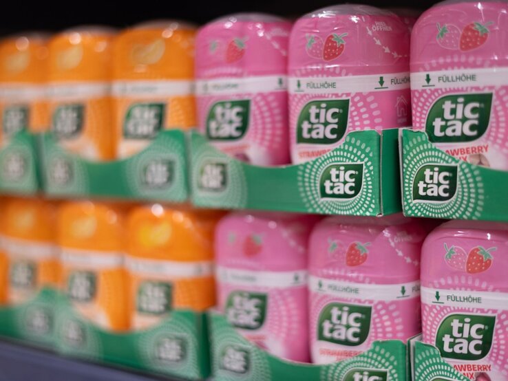 Regal mit orangefarbenen und rosa Tic Tac-Packungen in stapelbaren Pappträgern, im Vordergrund scharf fokussiert, im Hintergrund leicht verschwommen.