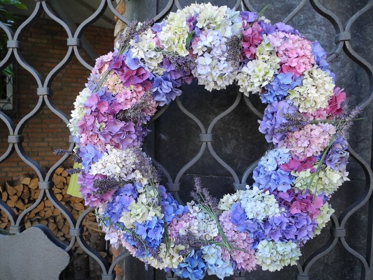 Hortensie Türkranz DIY Upcycling Romantischer Kranz aus farbenfrohen Hortensien und Lavendelzweigen, aufgehängt vor dunklem Gitterwerk, mit Holzscheiten im Hintergrund.