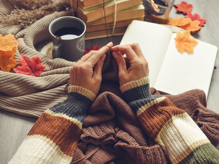 Hände einer Frau im gestreiften Pullover stricken braune Wolle, umgeben von Blättern, Büchern, einer Tasse Kaffee und Herbstdekoration.