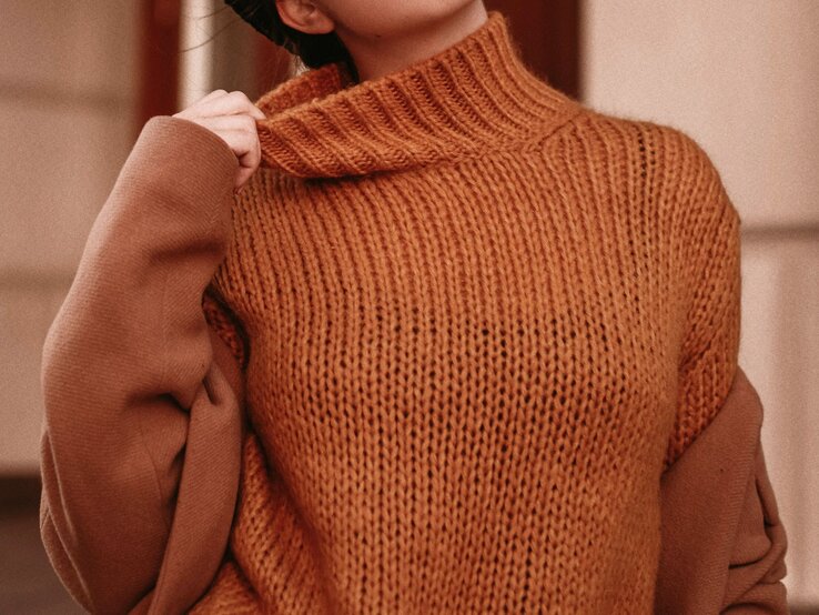 Frau zieht mit erhobener Hand den Kragen eines grob gestrickten orangebraunen Pullovers über einem Mantel auf beige-braunem Hintergrund.