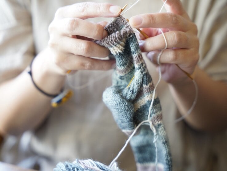 Hände halten Stricknadeln und arbeiten an einer gestrickten Socke in gedeckten Farben, Nahaufnahme mit weichem Hintergrund.
