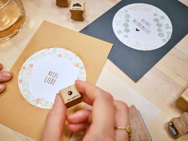 Gutschein Karte basteln Hand einer Frau mit goldfarbenem Ring stempelt liebevoll auf eine orange Karte mit der Aufschrift „Alles Liebe“ auf hellem Holztisch.