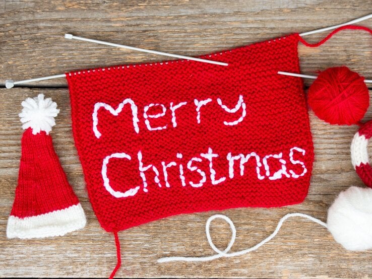 Rotes Strickstück mit weißem Schriftzug „Merry Christmas“ liegt mit Stricknadeln, Wolle und Mini-Weihnachtsmütze auf rustikalem Holz.