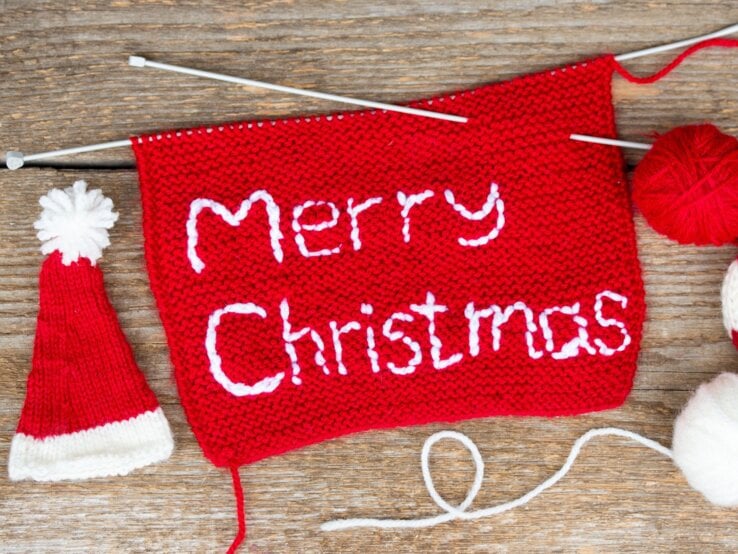 Rotes Strickstück mit weißem Schriftzug „Merry Christmas“ liegt mit Stricknadeln, Wolle und Mini-Weihnachtsmütze auf rustikalem Holz.