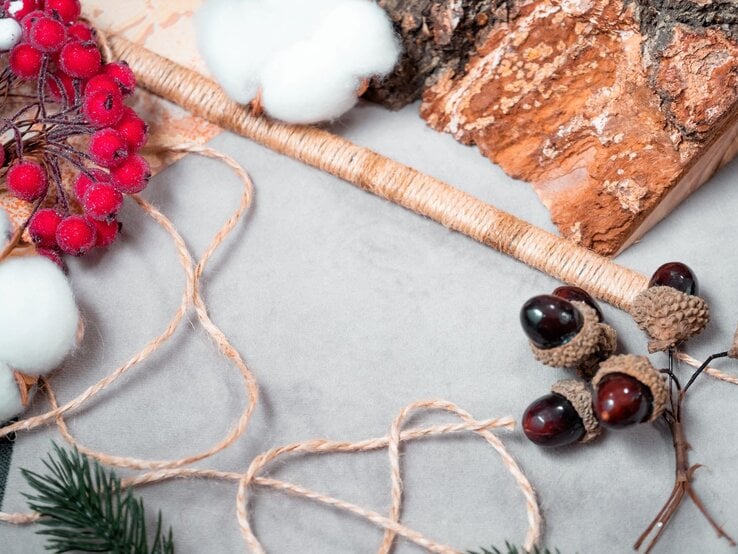 Naturmaterialien mit roten Beeren, Baumwolle, Eicheln und Juteschnur liegen dekorativ auf grauem Filzstoff und werden zu Halskette verarbeitet.