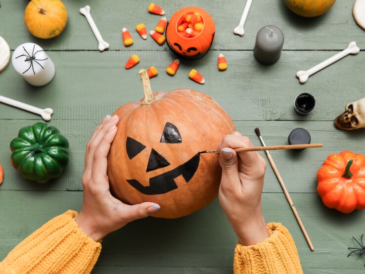 Zwei Hände bemalen einen Kürbis mit schwarzem Halloween-Gesicht, umgeben von Süßigkeiten und Deko auf grünem Holztisch.