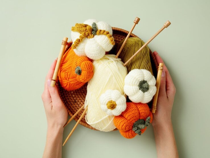 Zwei Hände halten einen Korb mit gestrickten Kürbissen, Wollknäueln in Herbstfarben und vier Holzstricknadeln.