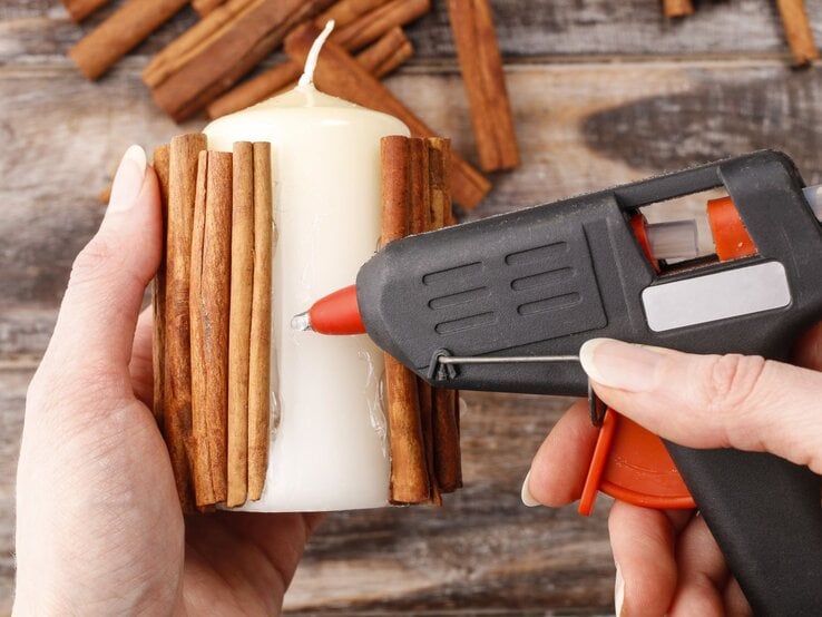 Zwei Hände kleben mit Heißklebepistole Zimtstangen auf eine weiße Kerze, im Hintergrund liegen weitere Zimtstangen.