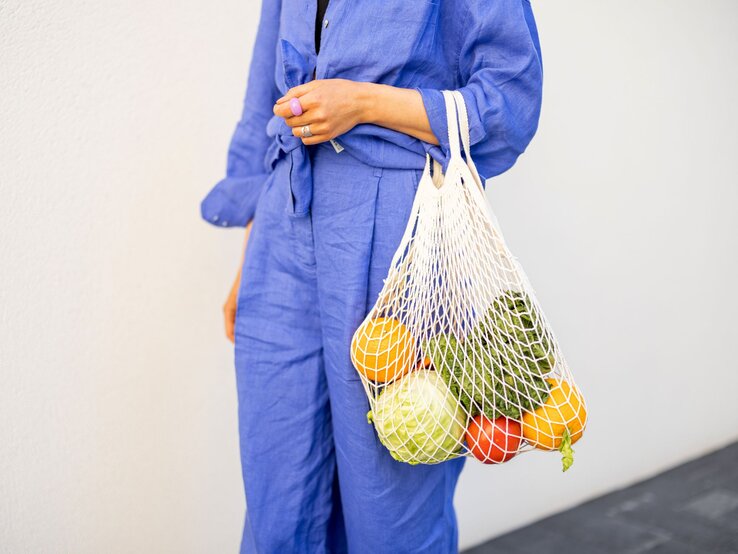 Detailaufnahme: Eine Hand mit Ring hält ein Einkaufsnetz mit Orangen, Tomate, Kohl und Salat, getragen zu einem blauen Outfit.