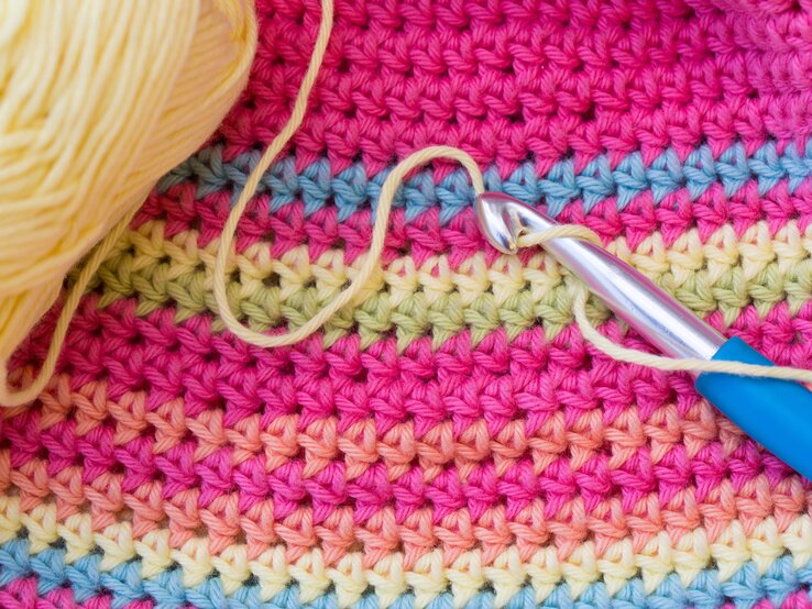 Pullover häkeln DIY Upcycling Detailaufnahme einer Häkelnadel mit gelbem Faden auf farbenfrohem Stoff in Rosa, Blau und Gelb, daneben ein Wollknäuel.