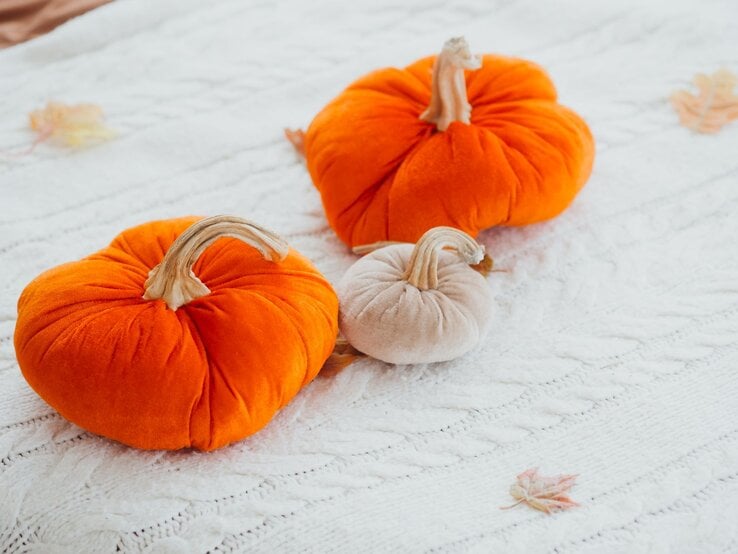 Zwei orangefarbene und ein beiger Zierkürbis aus Strumpfhosen gebastelt, liegen dekorativ auf einer weißen Strickdecke mit Herbstblättern.