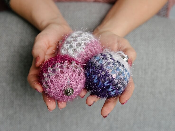 Nahaufnahme von Händen mit rot lackierten Nägeln, die drei gestrickte, glitzernde Weihnachtskugeln in Rosa, Lila und Blau halten.