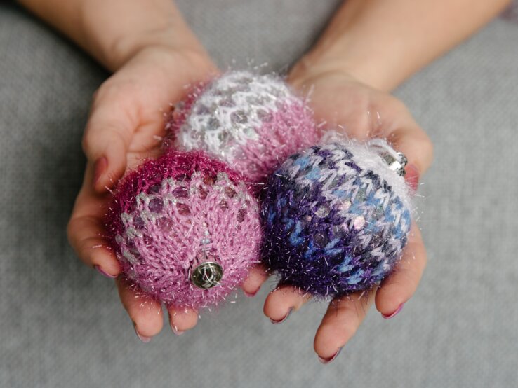 Nahaufnahme von Händen mit rot lackierten Nägeln, die drei gestrickte, glitzernde Weihnachtskugeln in Rosa, Lila und Blau halten.