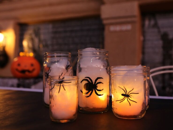 Vier Einmachgläser mit leuchtenden Kerzen und Spinnenmotiven stehen auf einem Tisch vor verschwommenem Halloween-Dekor.