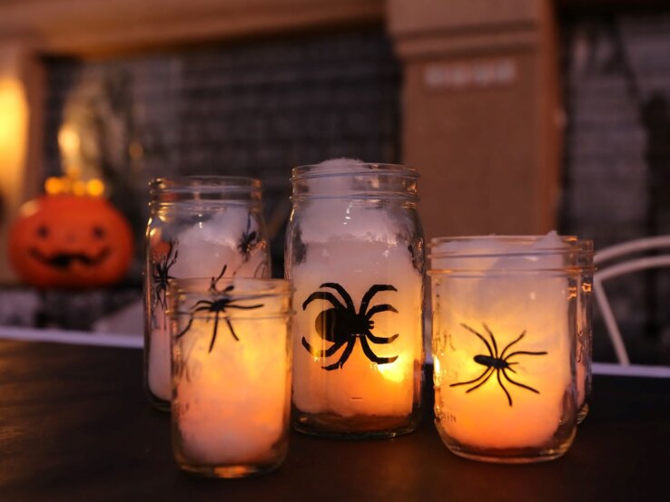 Halloween-Kerzen basteln  Vier Einmachgläser mit leuchtenden Kerzen und Spinnenmotiven stehen auf einem Tisch vor verschwommenem Halloween-Dekor.