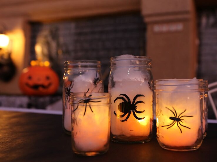 Halloween-Kerzen basteln  Vier Einmachgläser mit leuchtenden Kerzen und Spinnenmotiven stehen auf einem Tisch vor verschwommenem Halloween-Dekor.