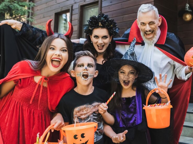 Verkleidete Familie posiert lachend vor gruselig geschmückter Haustür mit Kürbis, Eimern und DIY-Halloweenkostümen.