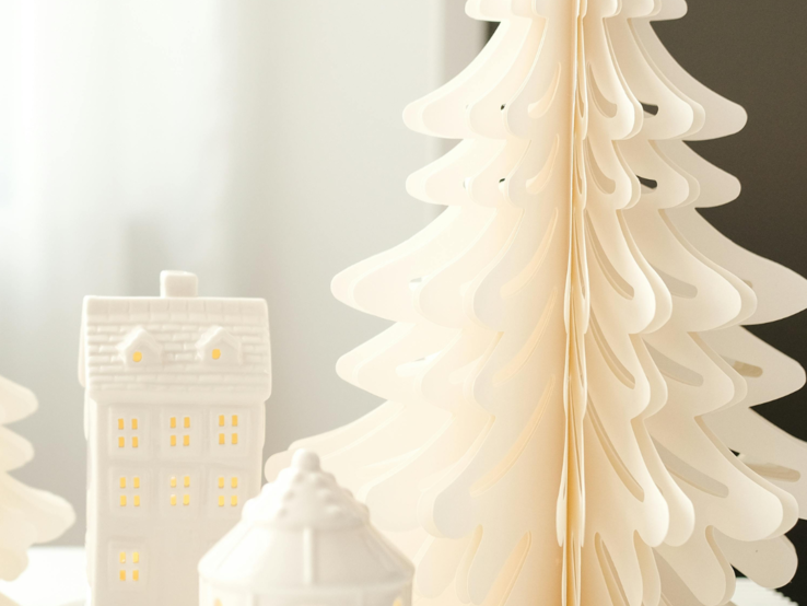 Faltbare Papierbäume in Weiß, Grün und Beige bilden mit leuchtenden Keramikhäuschen ein stimmungsvolles Winterensemble.
