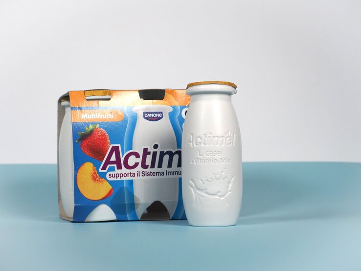 Basteln mit Actimel-Flaschen Halloweendeko  Eine weiße Actimel-Flasche mit goldener Versiegelung steht vor einem bunten Viererpack in Multifrucht-Design auf blauem Untergrund.
