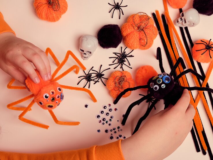 Pfeifenreiniger Halloween DIY Basteln Nahaufnahme von zwei Filzspinnen mit Pfeifenreinigern, Dekokürbisse, Schädel und viele Wackelaugen liegen verstreut auf hellbeigem Basteltisch.