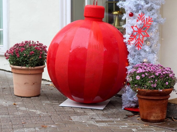 Riesige glänzend rote Weihnachtskugel steht zwischen zwei Blumentöpfen vor weißer Wand und geschmücktem Kunstbaum.