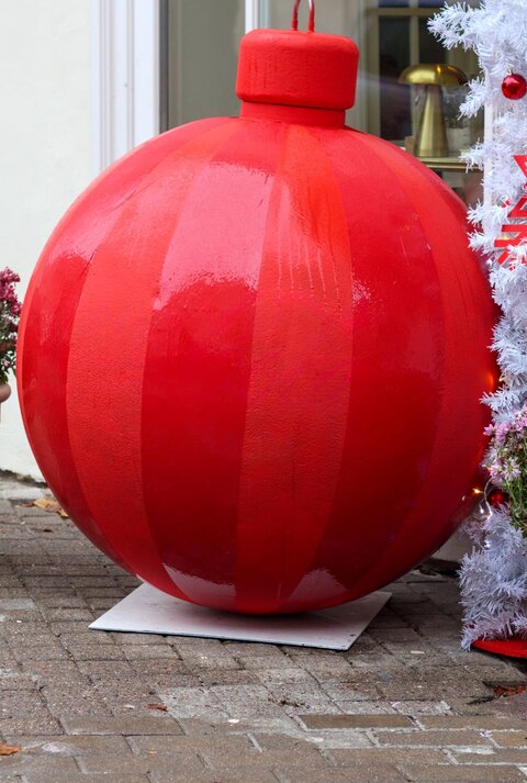 Riesige glänzend rote Weihnachtskugel steht zwischen zwei Blumentöpfen vor weißer Wand und geschmücktem Kunstbaum.