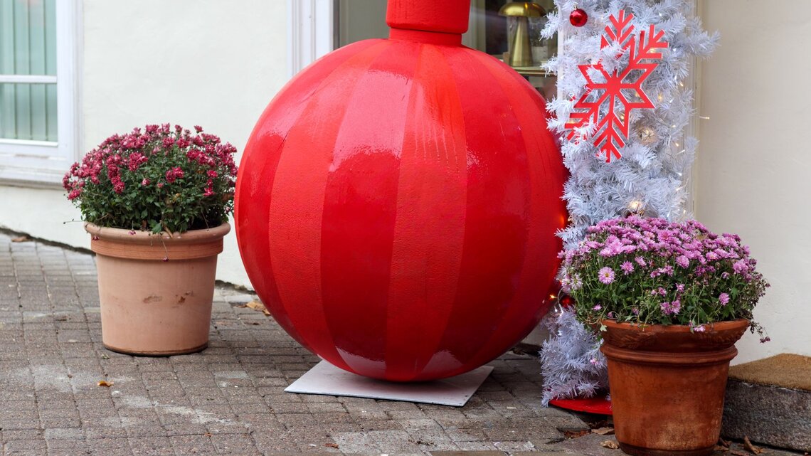 Riesige glänzend rote Weihnachtskugel steht zwischen zwei Blumentöpfen vor weißer Wand und geschmücktem Kunstbaum.
