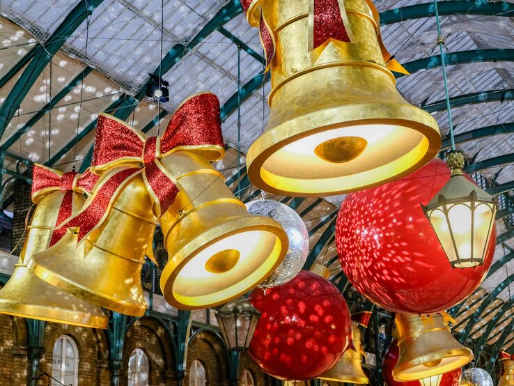 Riesige goldene Weihnachtsglocken mit roten Glitzerschleifen hängen leuchtend unter einem Glasdach mit Stahlbögen.