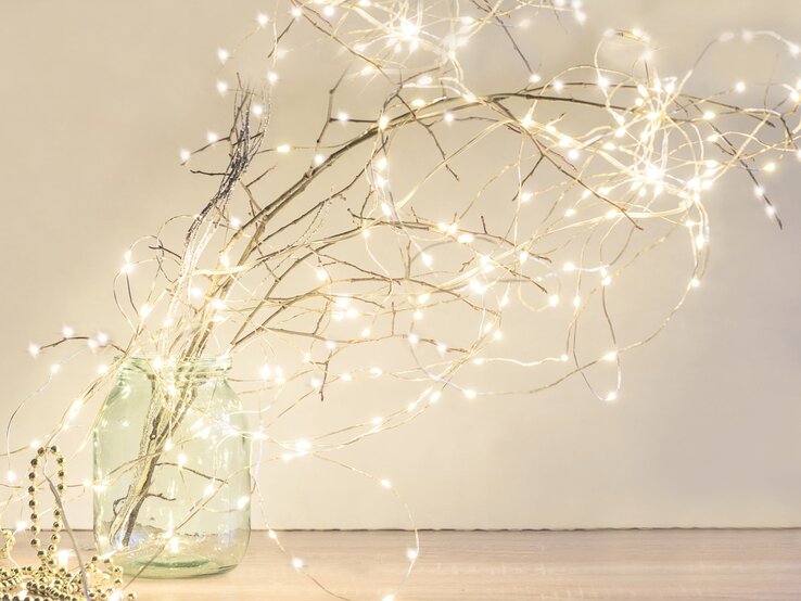 Ein bulliges glas mit Zweigen, die mit Lichterketten geschmückt sind, Hintergrund hell und die Lichterketten leuchten goldenweiss