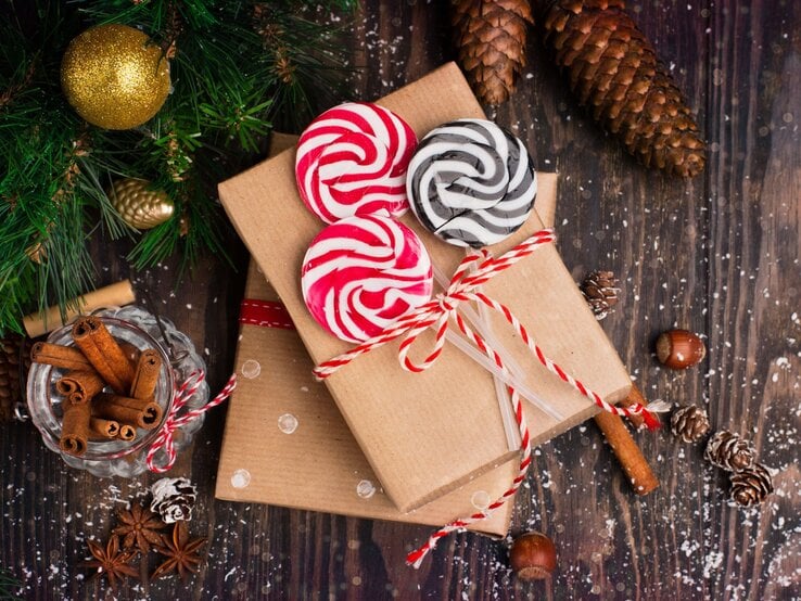 Zwei mit rotem Band dekorierte Gutschein-Geschenke, bunte Lollis und Zimtstangen liegen auf dunklem Holzboden, umgeben von Tannenzapfen.