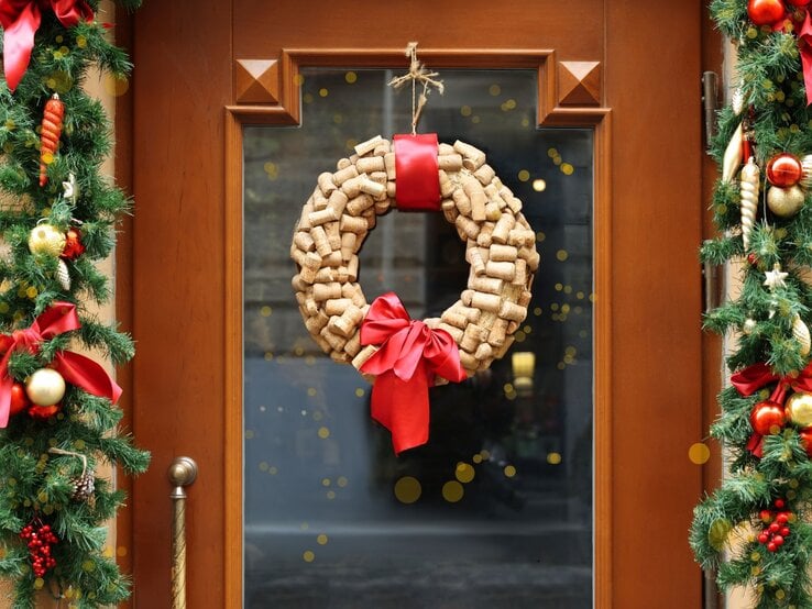 Weihnachtlich geschmückte Holztür mit Tannengirlanden und einem Kranz aus Korken, verziert mit roter Schleife.