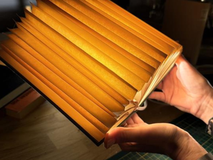 Mehrere kunstvoll gefaltete, warm leuchtende Buch-Lampen aus alten Seiten liegen auf einem Holztisch, eine wird gerade in Händen gehalten.