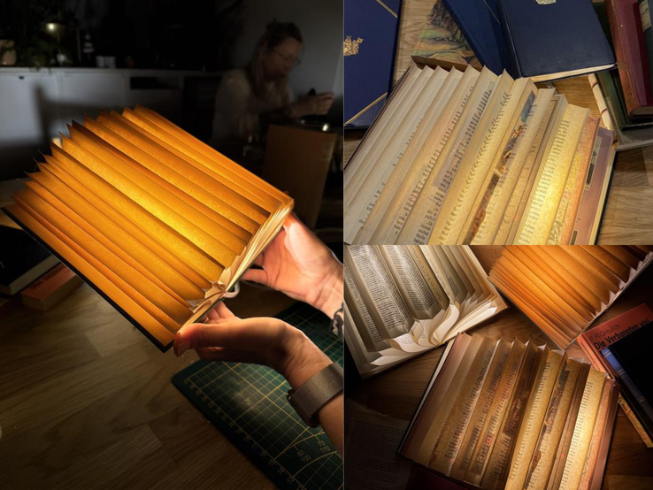 Mehrere kunstvoll gefaltete, warm leuchtende Buch-Lampen aus alten Seiten liegen auf einem Holztisch, eine wird gerade in Händen gehalten.