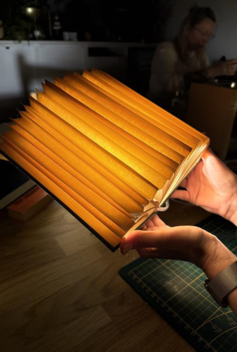 Mehrere kunstvoll gefaltete, warm leuchtende Buch-Lampen aus alten Seiten liegen auf einem Holztisch, eine wird gerade in Händen gehalten.