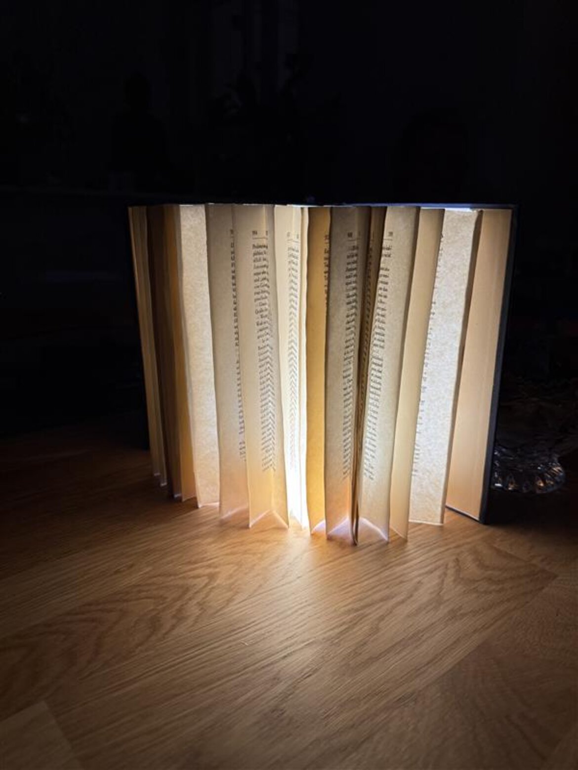 Eine aufgefächerte Buch-Lampe mit gelblich leuchtenden Seiten steht auf einem Holztisch in dunkler Umgebung. | © Madeleine Liedke 
