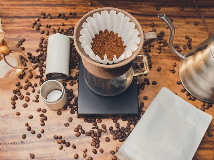 Frisch gemahlener Kaffee liegt im Papierfilter eines Handfilters, umgeben von Bohnen, Mühle und Kanne auf Holztisch.
