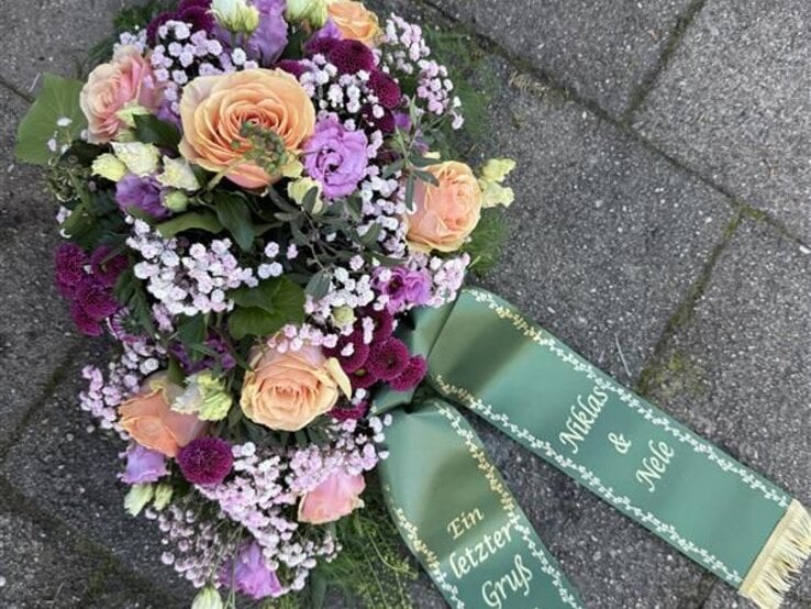 Ein buntes Grabgesteck mit Rosen, Nelken und Schleierkraut liegt mit grünem Schleifenband auf grauen Pflastersteinen.