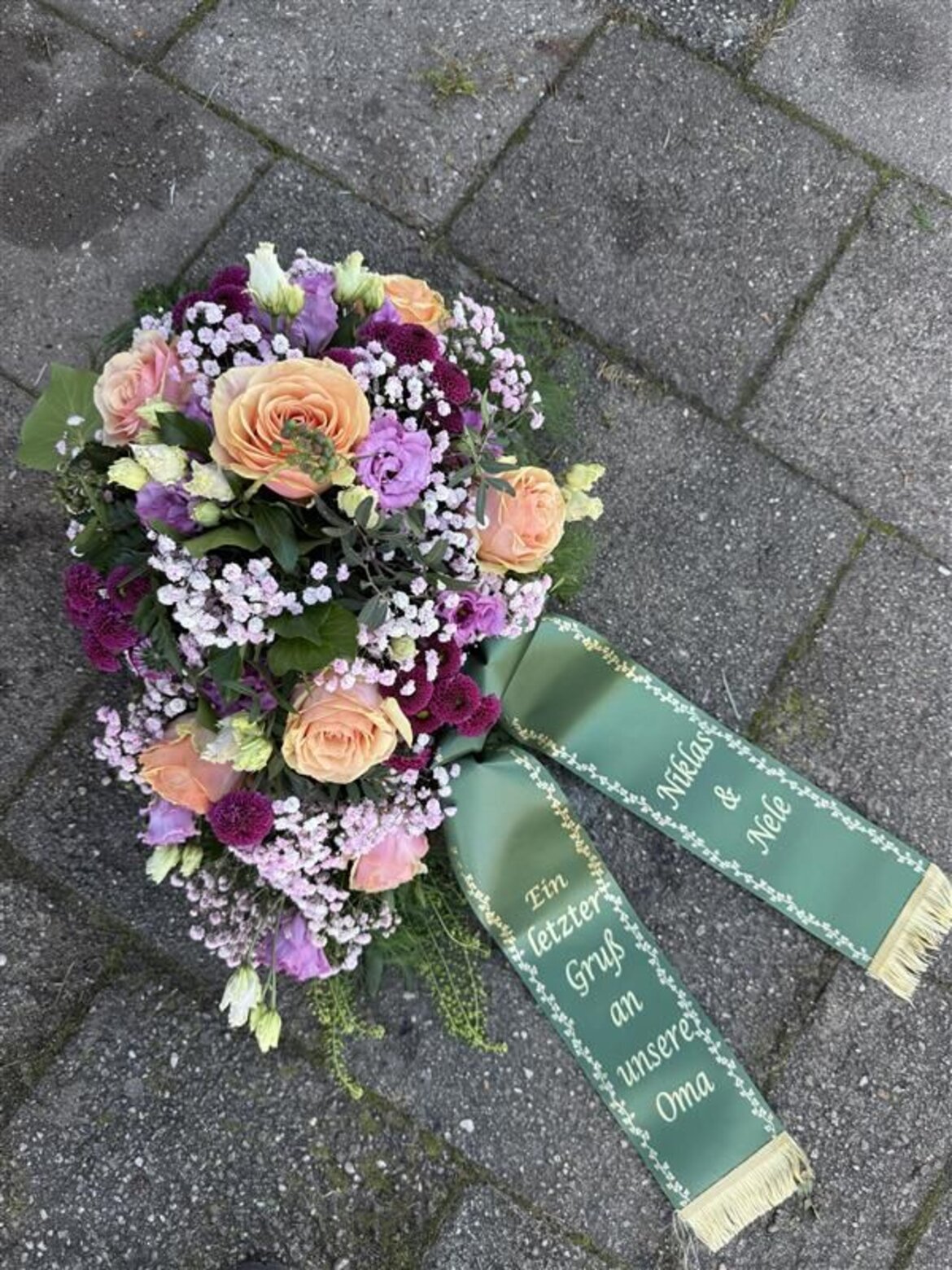 Ein buntes Grabgesteck mit Rosen, Nelken und Schleierkraut liegt mit grünem Schleifenband auf grauen Pflastersteinen. | © Madeleine Liedke 