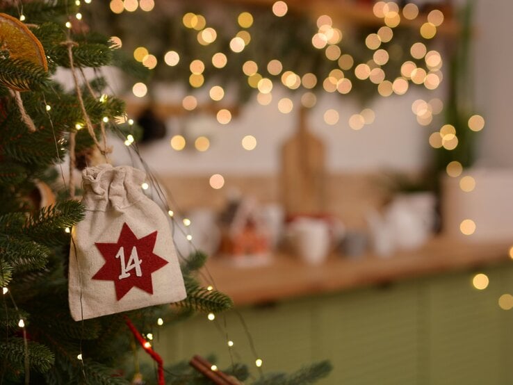 Adventskalender-Food: Ein kleiner Leinensack mit rotem Stern und Zahl 14 hängt an einem Tannenzweig, umgeben von warmen Lichterketten.