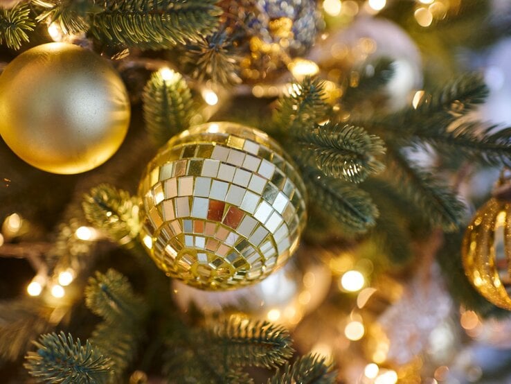 Eine goldene Christbaumkugel in Discokugel-Optik spiegelt Lichter zwischen Tannenzweigen auf einem festlich geschmückten Baum.
