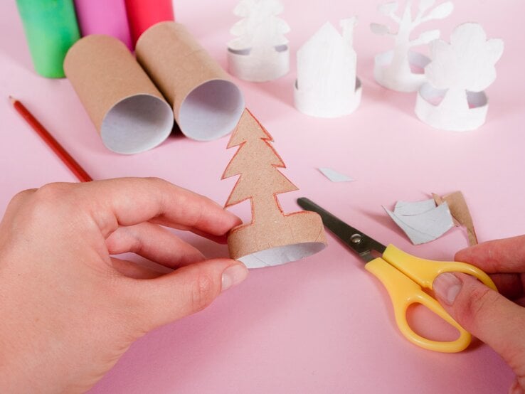 Zwei Hände basteln aus einer Klopapierrolle einen Tannenbaum-Teelichthalter, im Hintergrund weitere Figuren aus Karton.