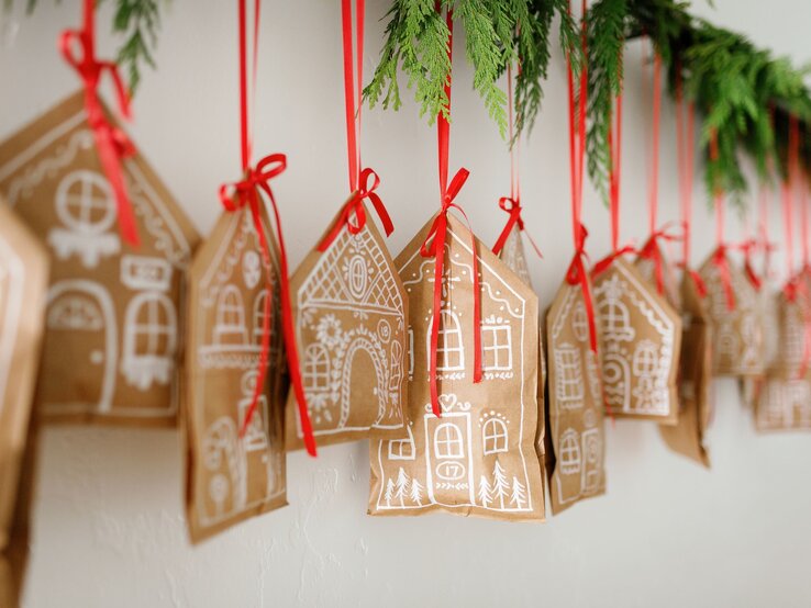 Zwanzig kleine Lebkuchenhäuschen mit weißem Muster hängen als Girlande an roten Bändern unter Tannenzweigen.